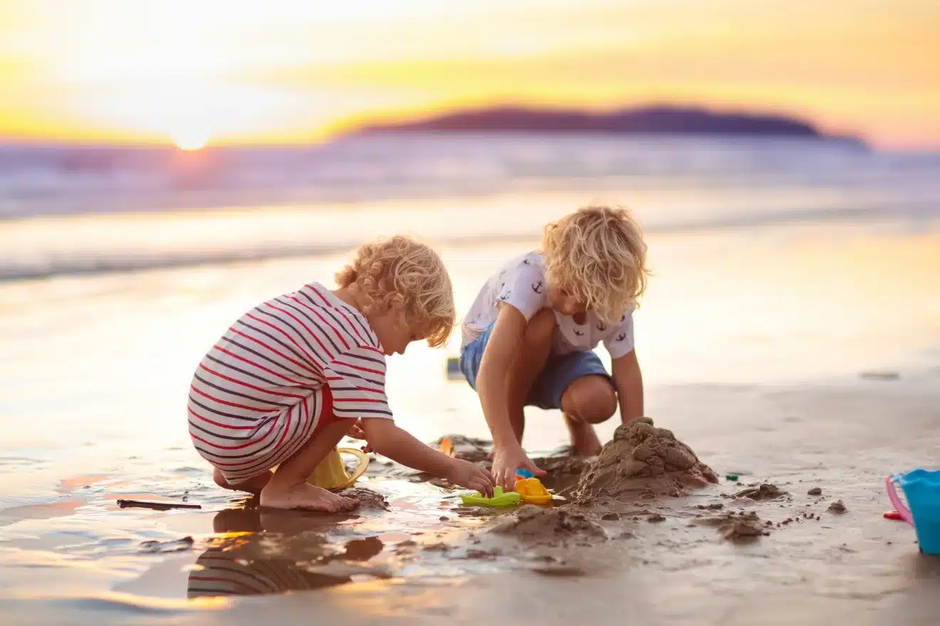 Two children with blond hair playing with toys and building a sandcastle on a beach at sunset.