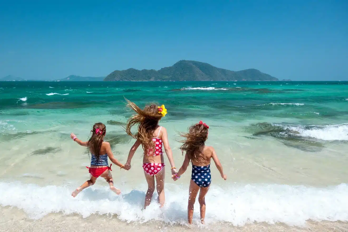 Three children in colorful swimsuits play at the edge of the ocean, holding hands and facing the waves. An island and clear blue sky are visible in the background.