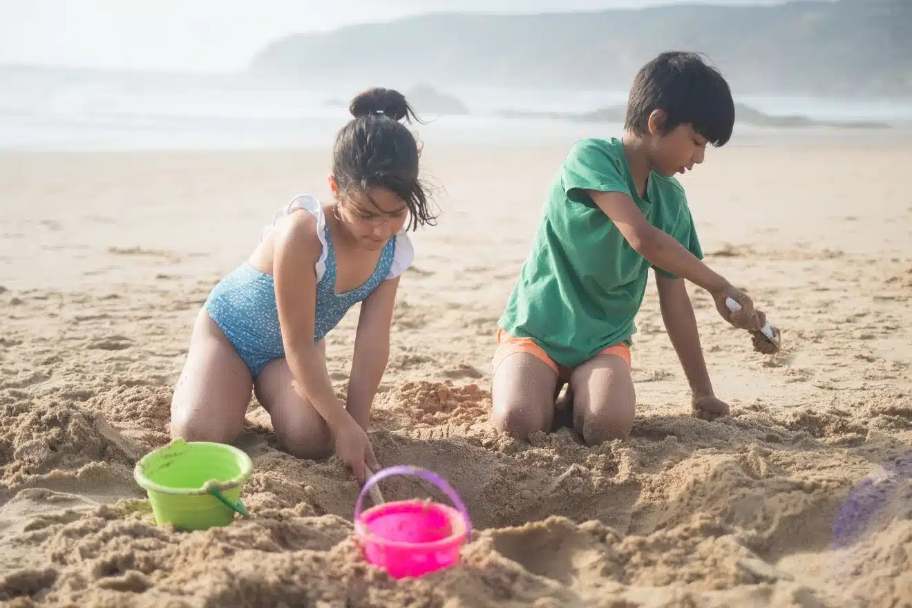Two children play in the sand on a beach, scooping sand with their hands and plastic buckets. The ocean and hills are visible in the background.