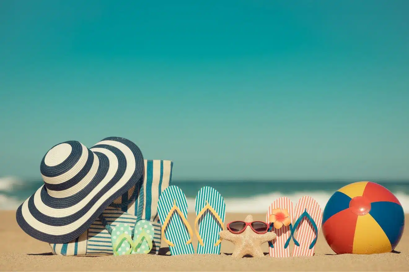 Beach items on sand: a striped hat, towel, and bag, four pairs of flip-flops, a starfish with sunglasses, and a beach ball. Ocean and clear sky in the background.