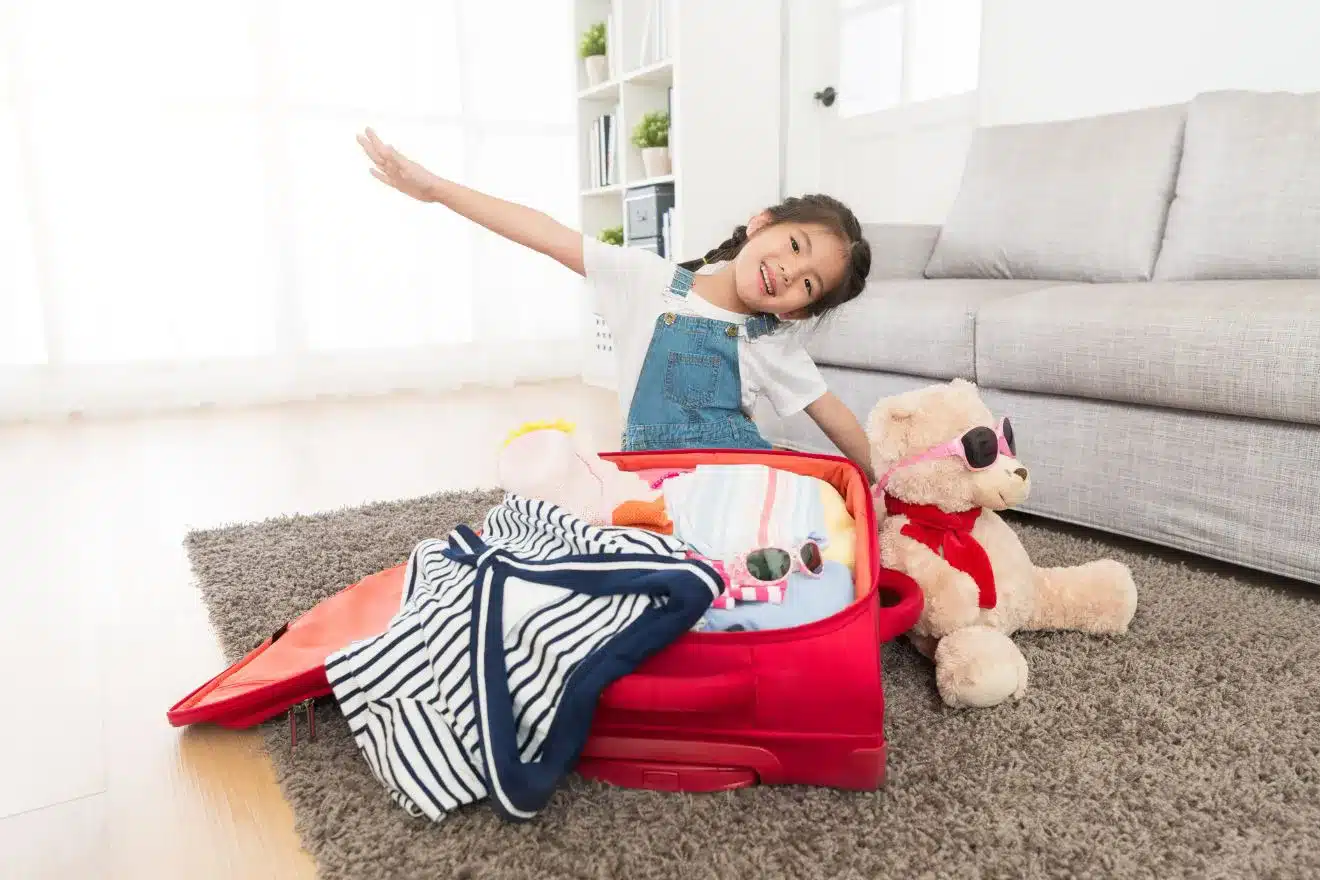 A young girl smiles while sitting beside a suitcase filled with clothes and a stuffed bear wearing sunglasses.