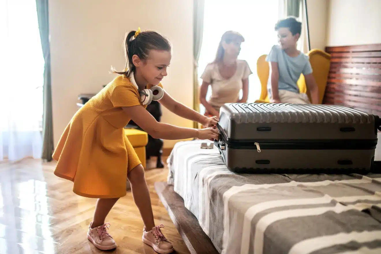 A young girl in a yellow dress is opening a suitcase on a bed while a woman and a boy are sitting in the background in a hotel room. kids packing list