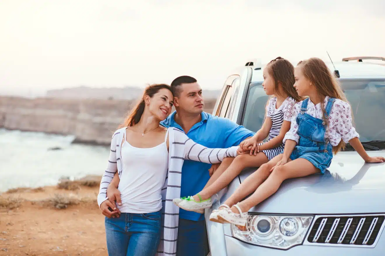 A family of four stands by an SUV near a coastal cliff. The parents embrace each other, while the two young girls sit on the car's hood, smiling and interacting.