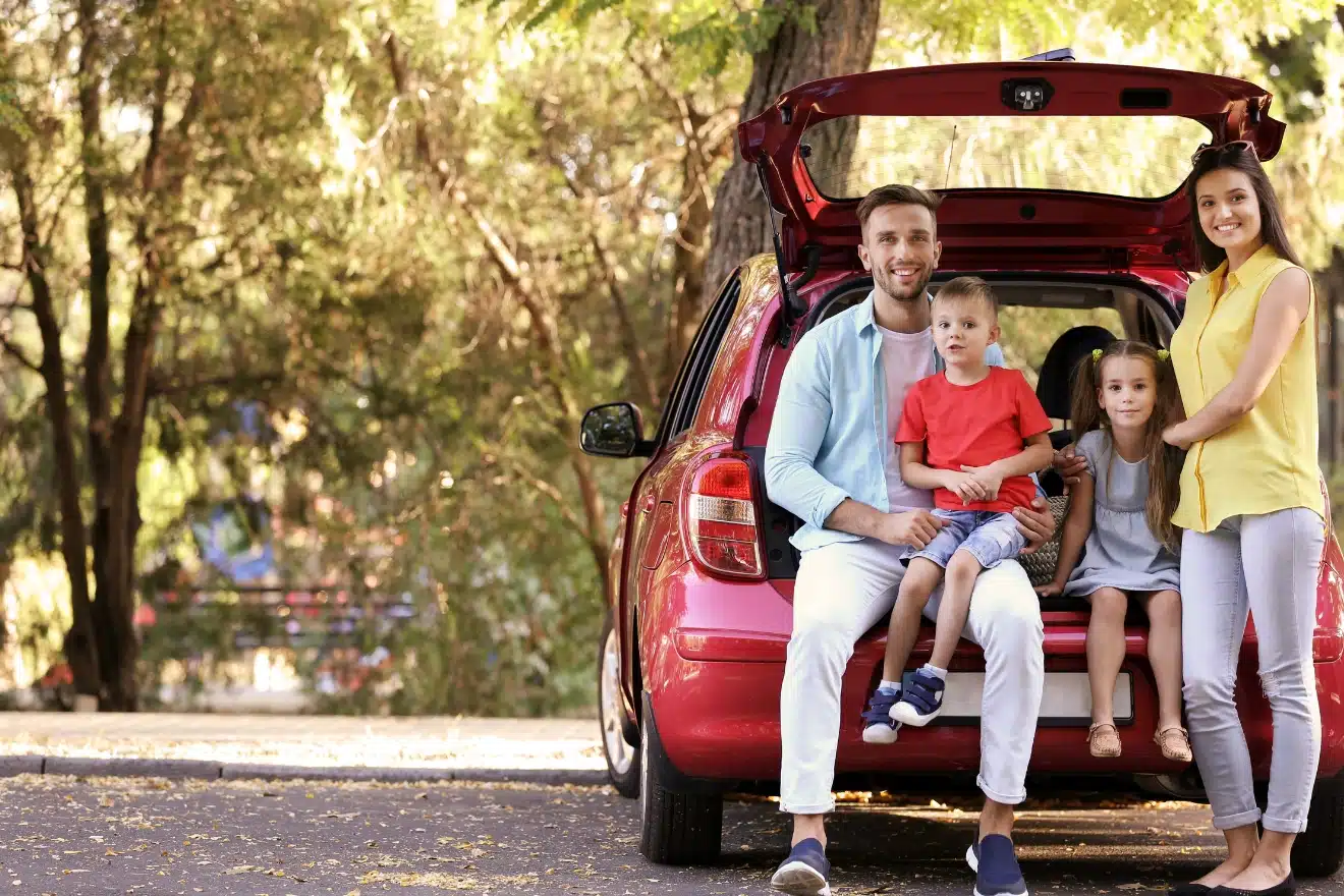 A family of four sits and stands by the open trunk of a red car in a wooded area. The two children, kept entertained with a road trip maze, sit on the edge of the trunk while the parents stand nearby.