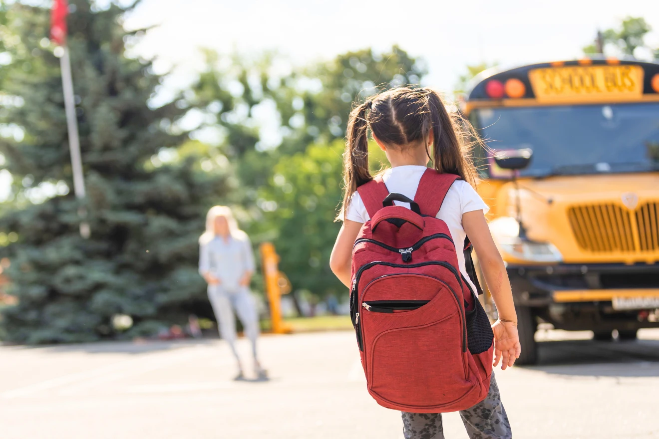A young girl with a red backpack stands facing a yellow school bus, with another person in the background.