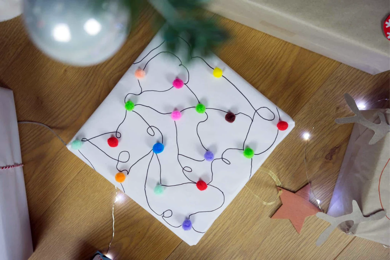 A wrapped gift is decorated with a winding black line and assorted colorful pom-poms. A small model house and other decorative items are in the background.