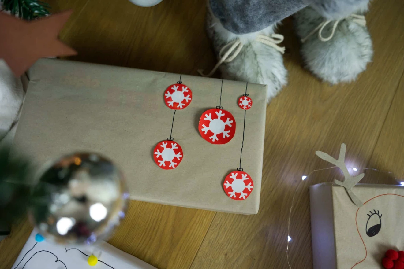 A wrapped gift decorated with red and white snowflake ornaments sitting on a wooden floor. Partially visible plush feet are beside the gift. Another wrapped gift with a reindeer drawing is nearby.