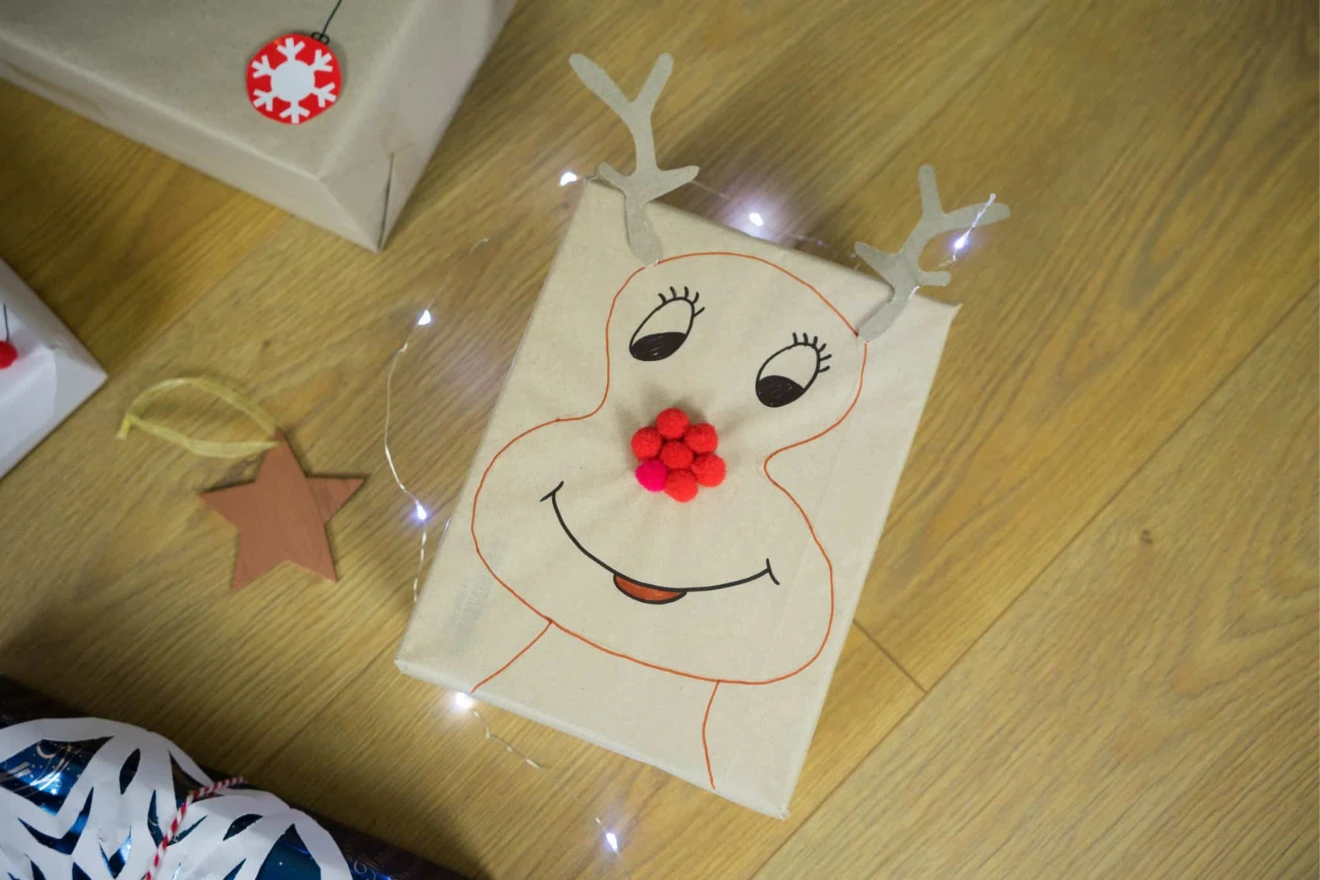A gift wrapped in paper with a reindeer face, featuring a red pom-pom nose and antlers, is surrounded by a string of small lights and a wooden star ornament on a wooden floor—perfect gift wrapping ideas for kids.