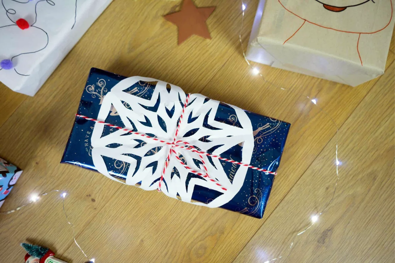 A wrapped gift with a blue wrapping paper and a white paper snowflake decoration on top, tied with a red and white string, placed on a wooden floor among other wrapped presents.