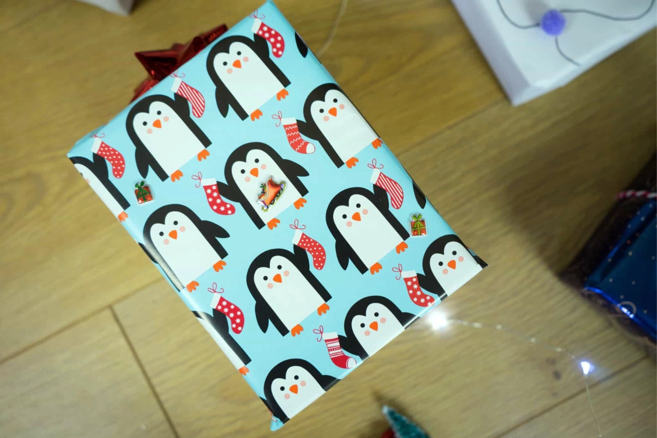 A gift wrapped in blue paper with penguin, stocking, and gift box designs sits on a wooden floor.