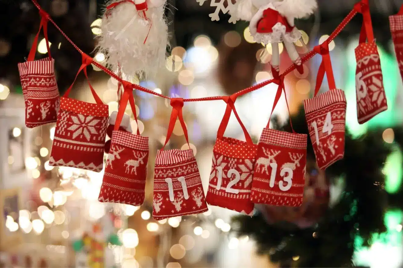 Red and white knitted advent calendar bags with numbers 11 to 14 hang on a string, surrounded by festive decorations.