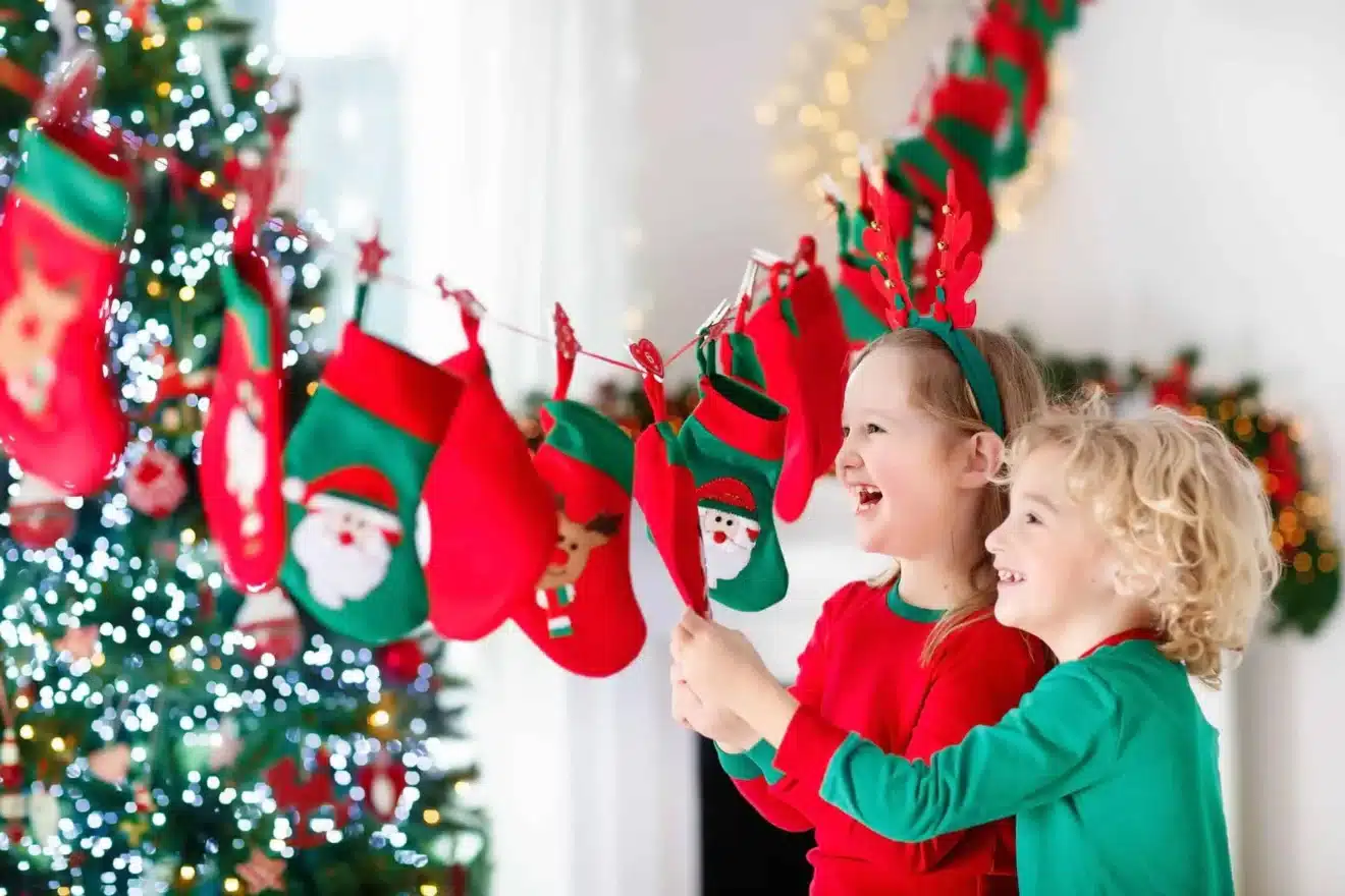 Two children in festive pajamas smile and look at Christmas stockings hanging on a garland, with a decorated Christmas tree in the background.