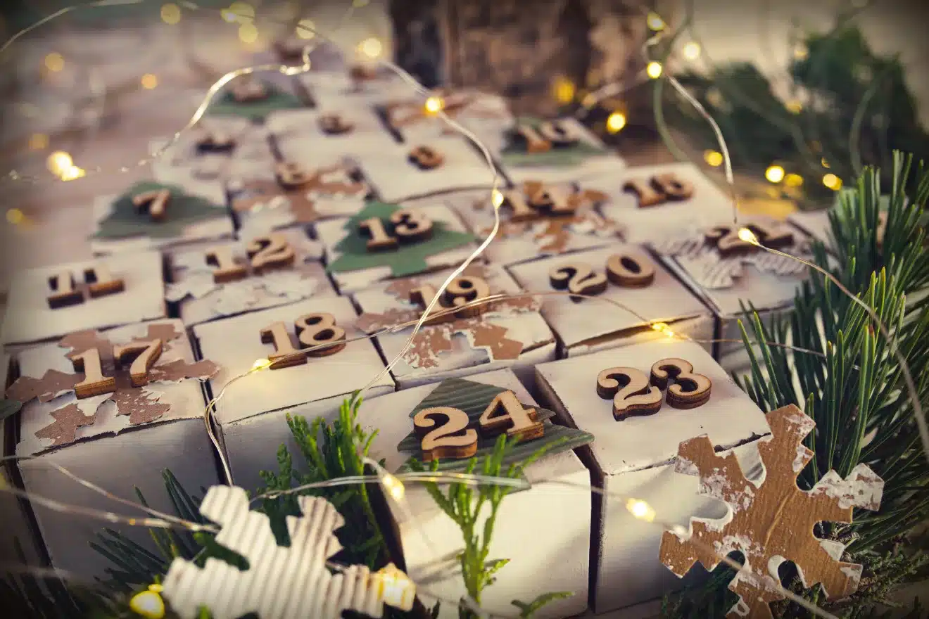 A close-up of a homemade advent calendar made of small numbered boxes, surrounded by fairy lights, greenery, and decorative snowflakes.