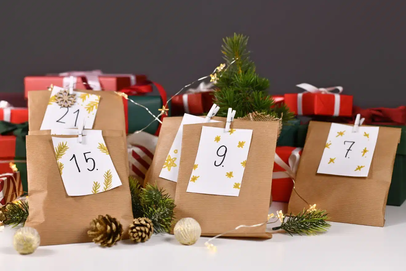 Brown paper bags with numbers 7, 9, 15, and 21, decorated with pine leaves and stars, placed among red and green gift boxes, pine cones, and fairy lights.