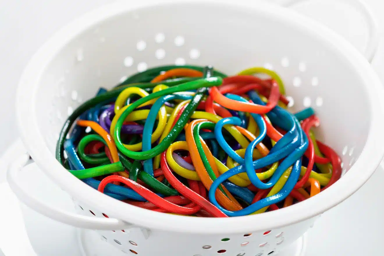 A white colander filled with cooked spaghetti noodles dyed in various bright colors, including red, blue, green, yellow, and orange.
