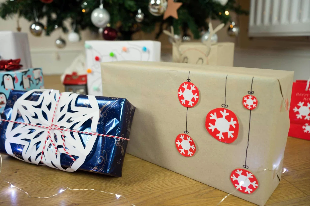 Two wrapped gifts sit on a table near a decorated Christmas tree. One is in blue paper with a white snowflake cutout, and the other is in plain brown paper with red and white ornament decorations.