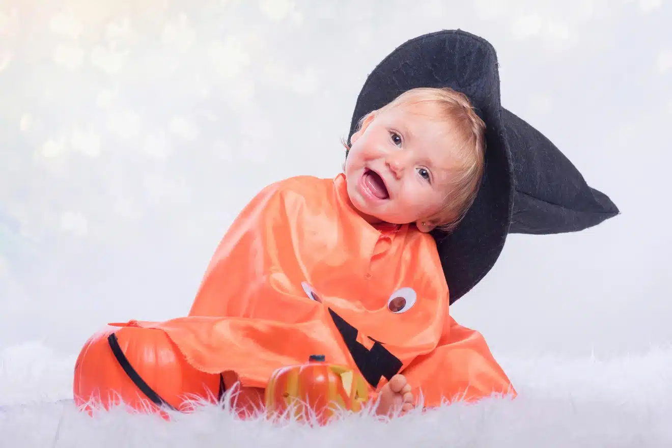 A young child wearing an orange pumpkin costume and a black hat sits on a white surface, surrounded by small pumpkins.