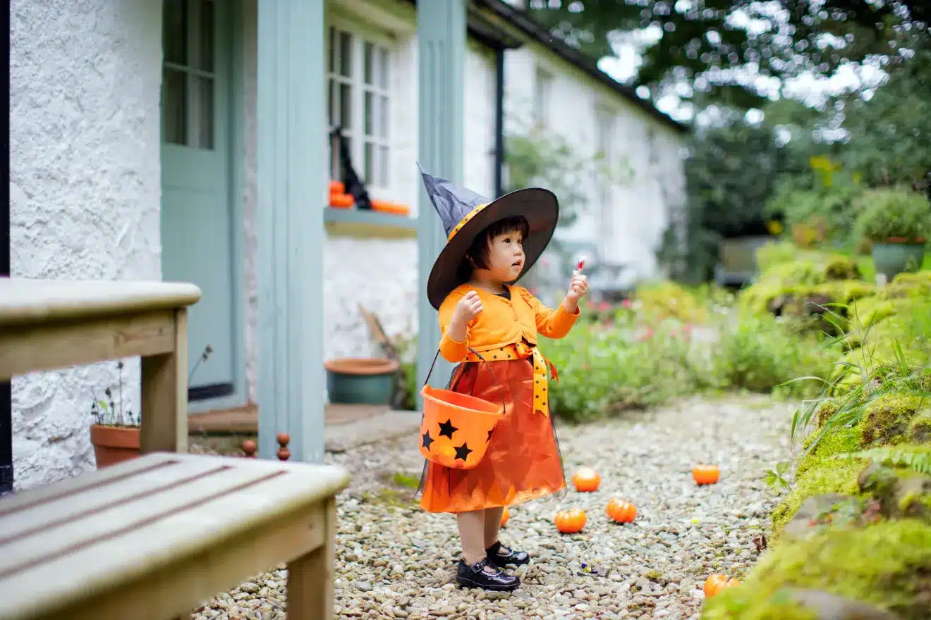 A young child dressed in a witch costume, holding a lollipop, and standing in a garden with pumpkins scattered around. The child wears a pointed hat and carries a pumpkin-shaped bag.