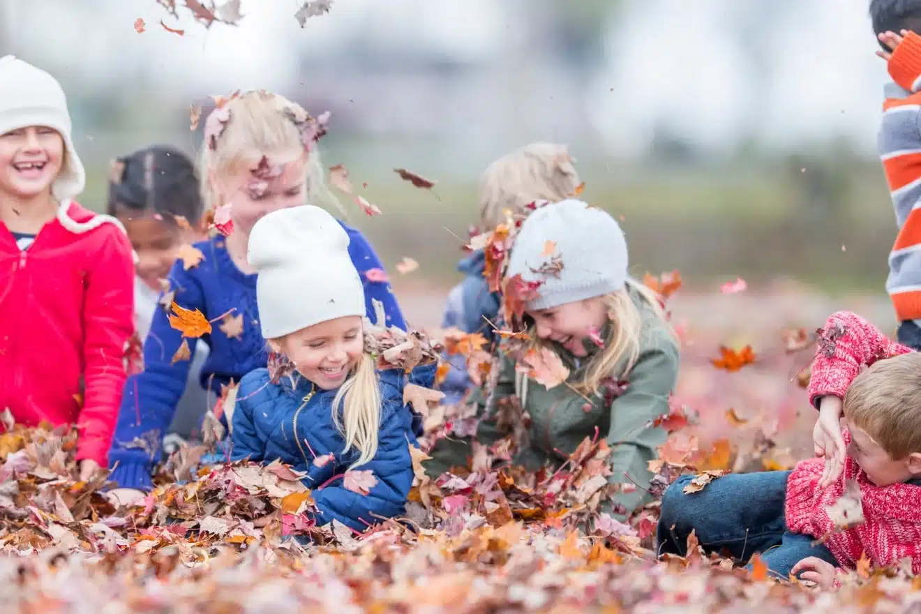 Children playing joyfully in a pile of autumn leaves, wearing colorful jackets and hats.