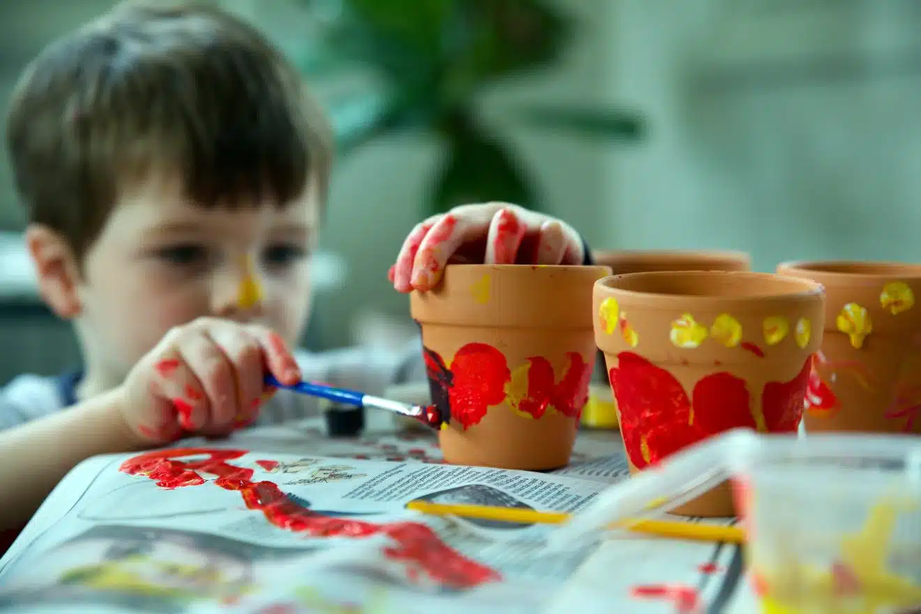 A child paints clay pots with red and yellow designs, using a paintbrush and their hands.