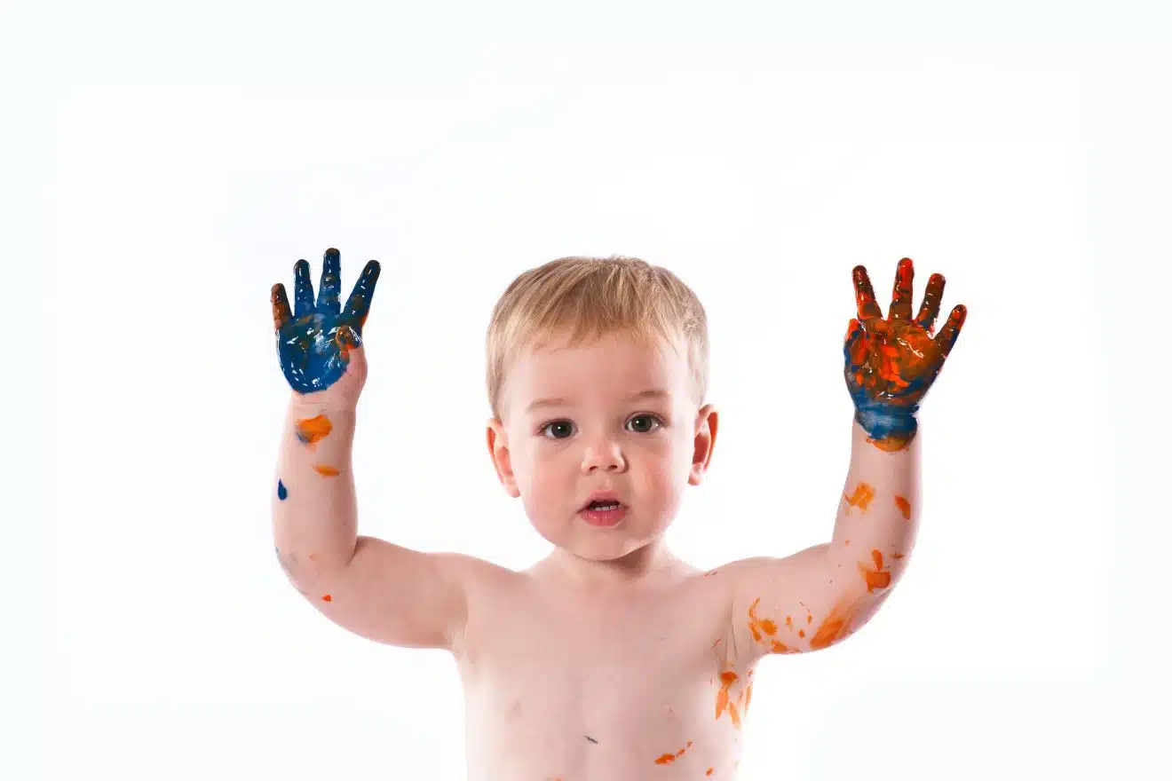 A child with paint-covered hands raised, featuring blue and orange colors, against a plain background.