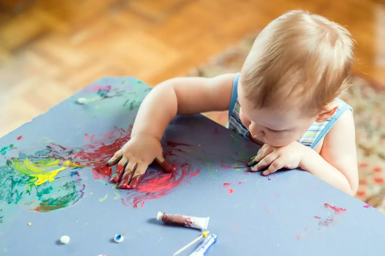 A toddler finger-paints on a table with colorful swirls of red, yellow, and green. Open paint tubes are nearby.