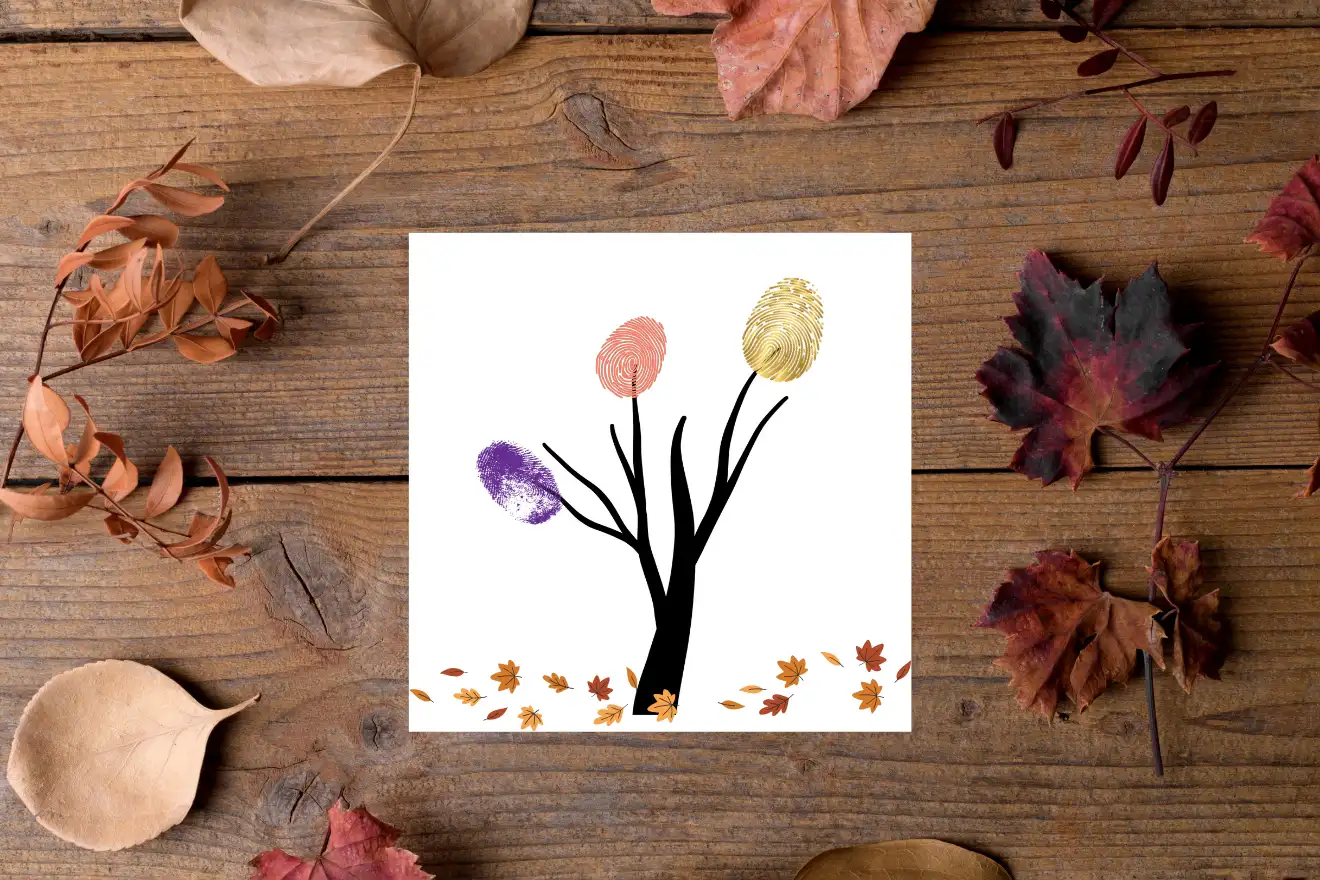 A drawing of a tree with fingerprint leaves in various colors on white paper, surrounded by autumn leaves on a wooden surface.