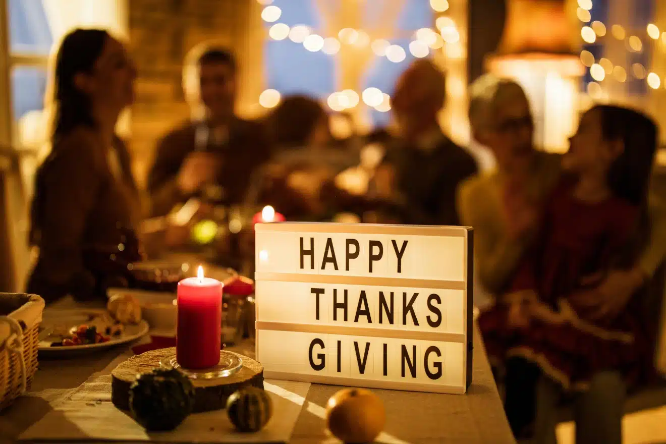 A Thanksgiving gathering with a "Happy Thanksgiving" sign in the foreground, surrounded by a candle and food items. People are seated and chatting in a warmly lit room.