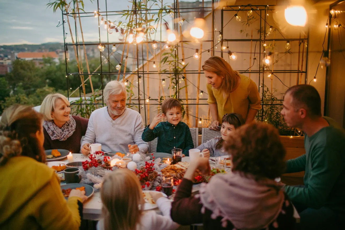 A group of people sit around a table outdoors, enjoying a meal together. The setting is decorated with string lights and greenery.