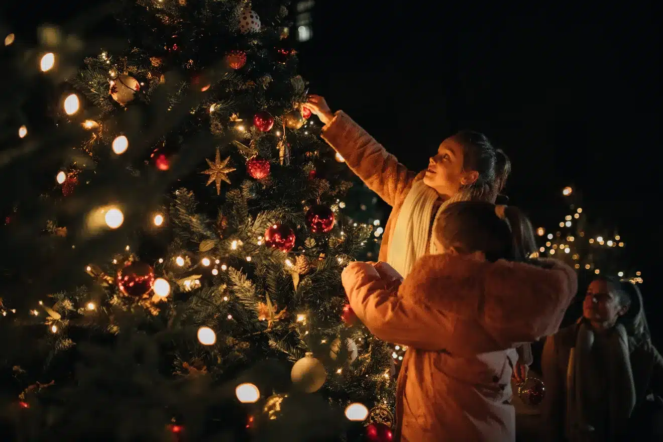 A person and child decorate an outdoor Christmas tree with lights and ornaments at night. Another person stands nearby, observing.