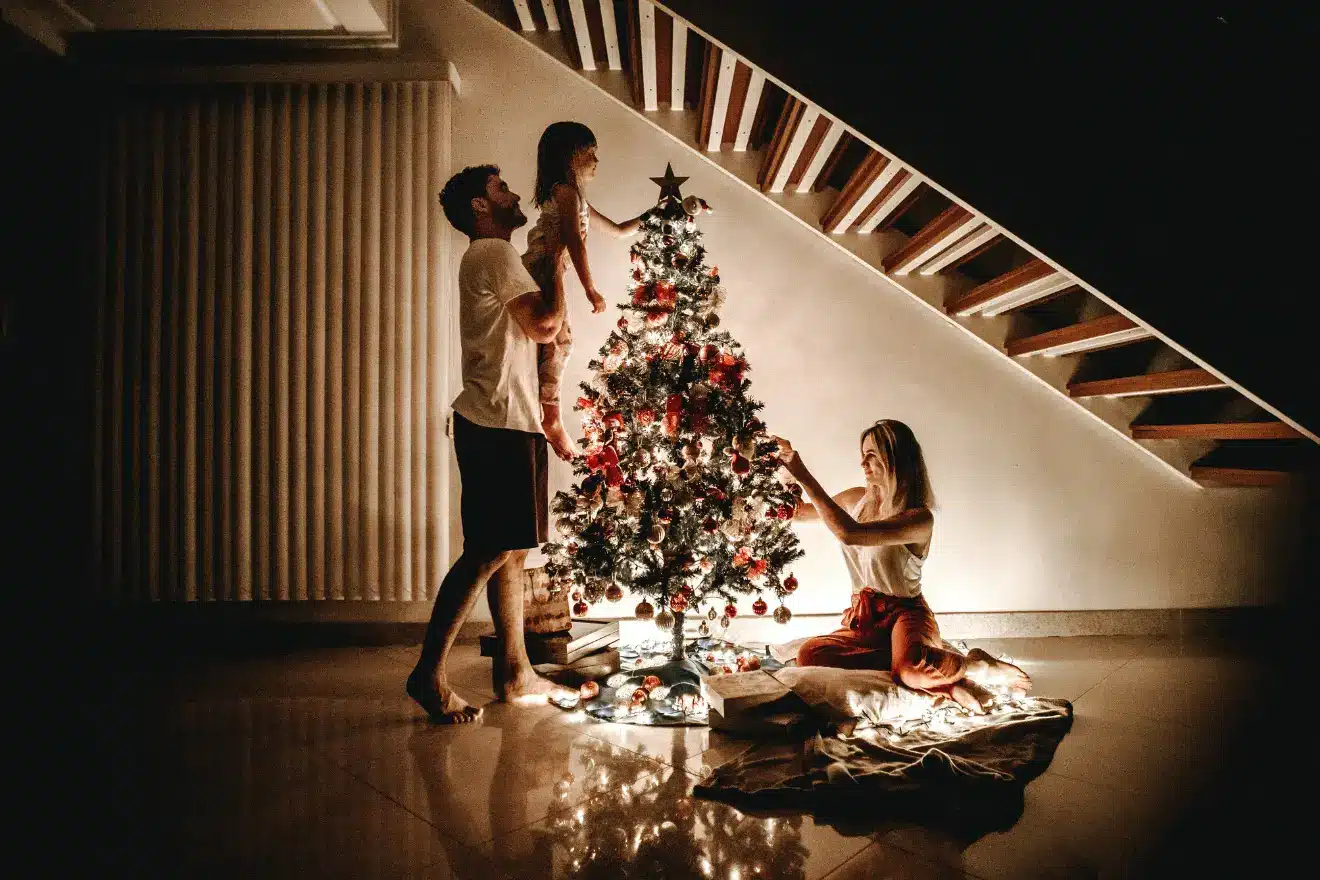 A family decorates a Christmas tree with lights and ornaments in a dimly lit room, next to a staircase.