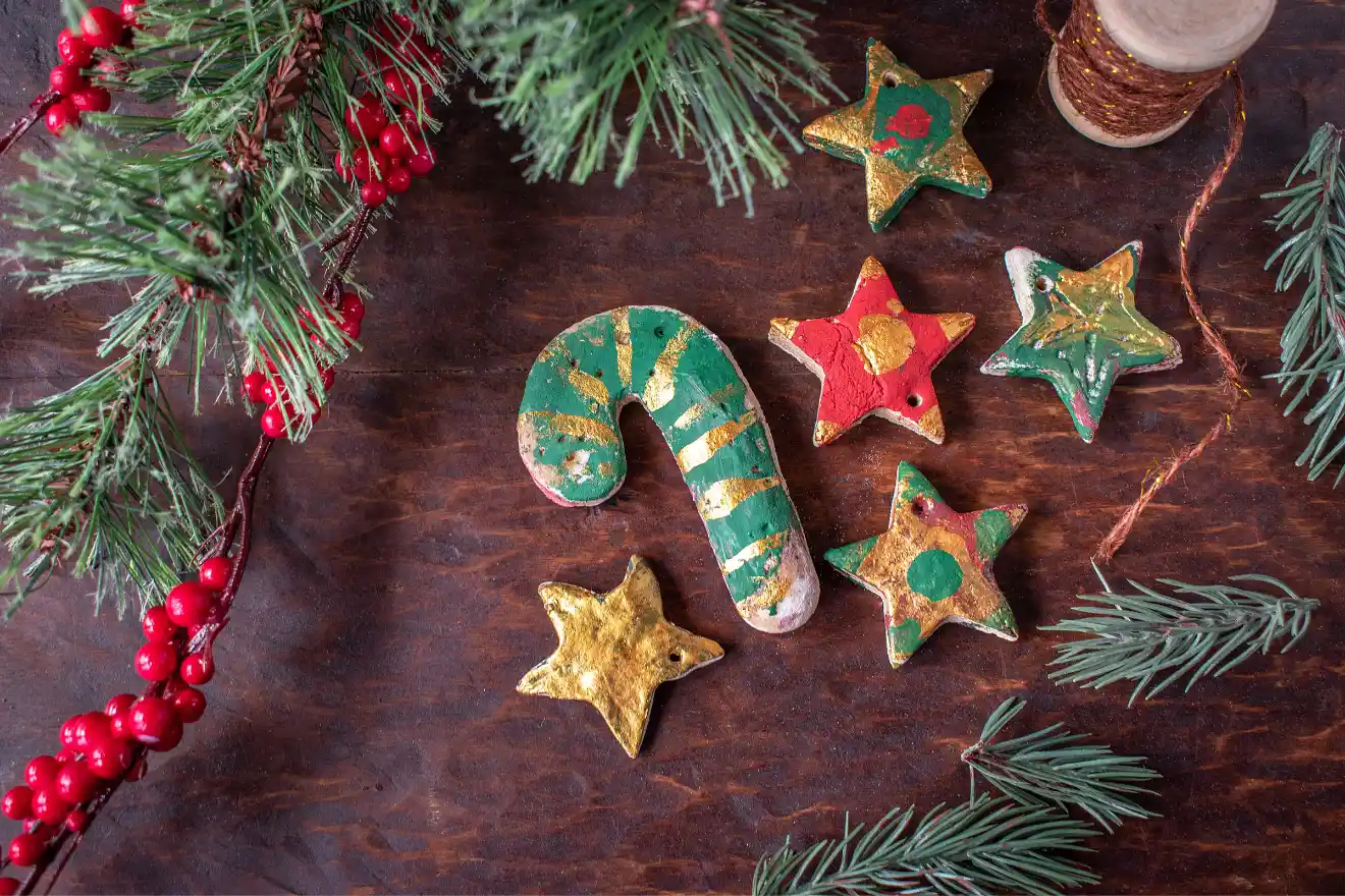 Wooden decorations including a green and gold candy cane and colorful stars are arranged on a wooden surface, surrounded by sprigs of evergreen and red berries.