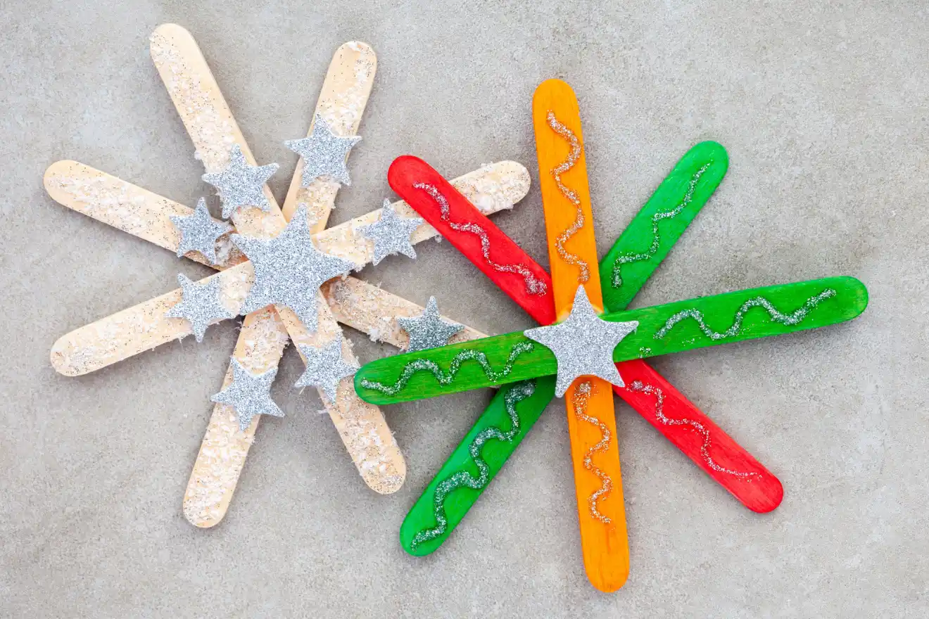 Craft stars made from colored popsicle sticks and silver glitter stars are arranged on a gray background. Two stars are multicolored, and one is beige.