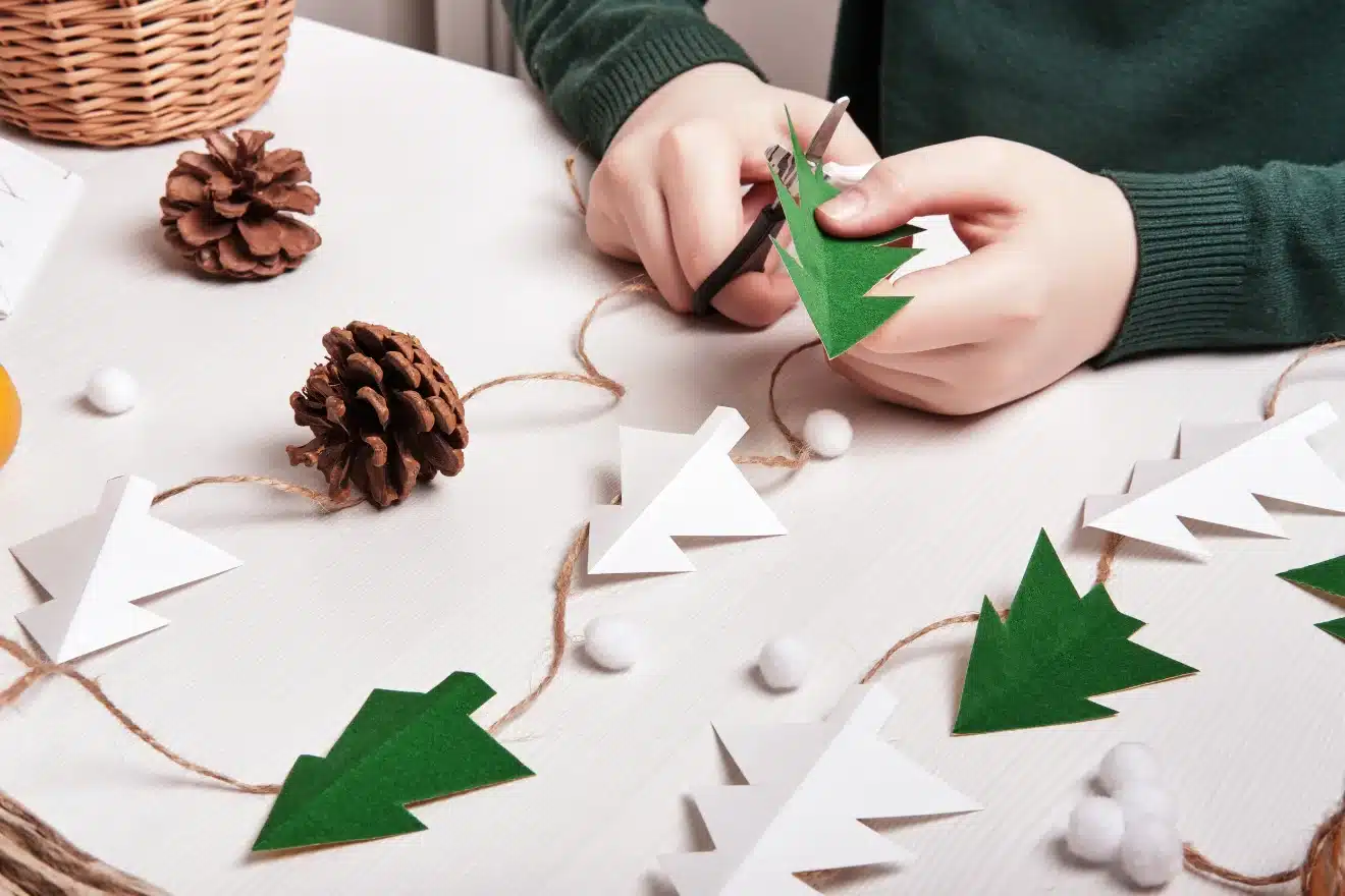 Person cutting green paper trees on a table with pine cones, white pom-poms, and paper tree garlands.