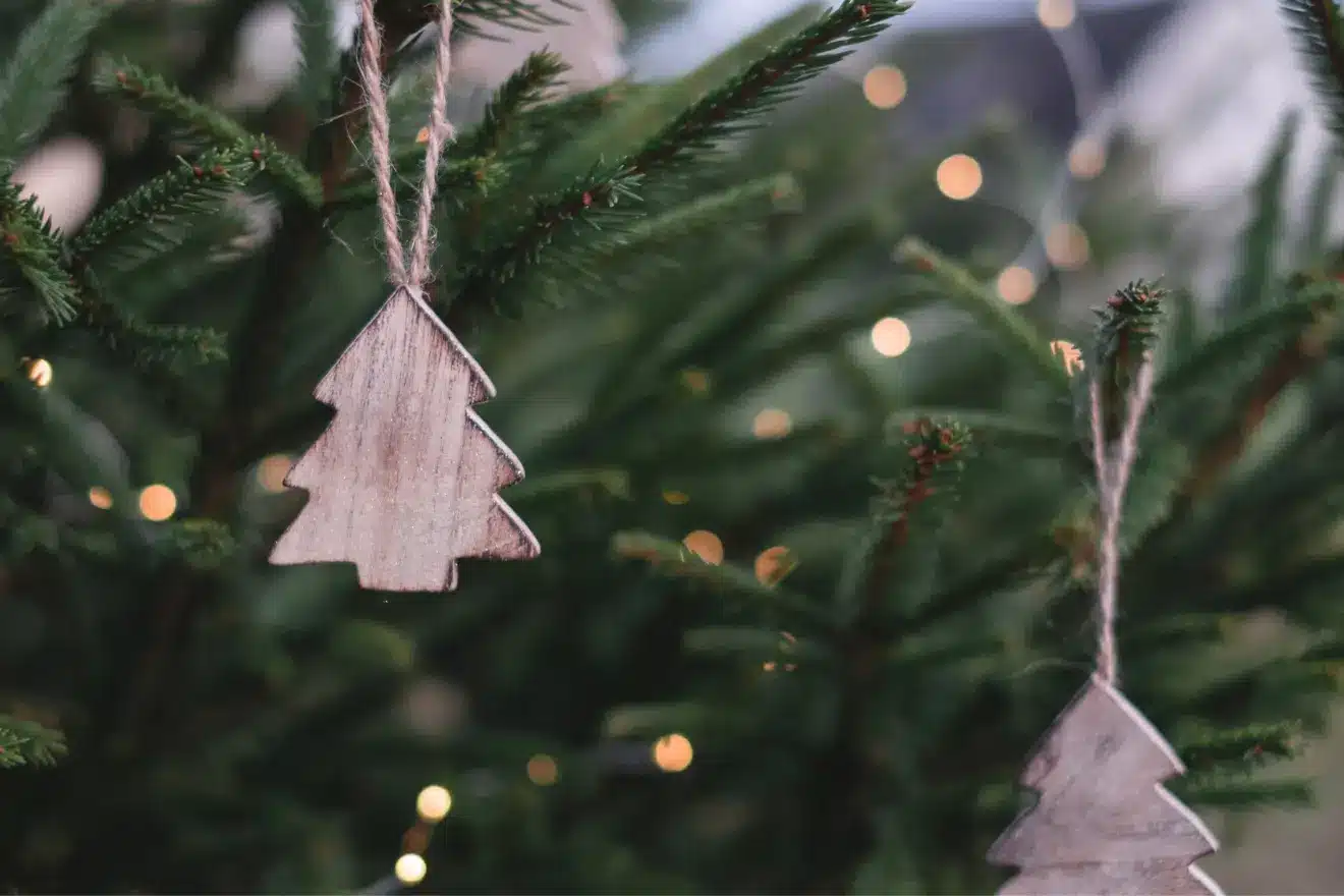Wooden tree-shaped ornaments hang on a pine tree, with blurred lights in the background.