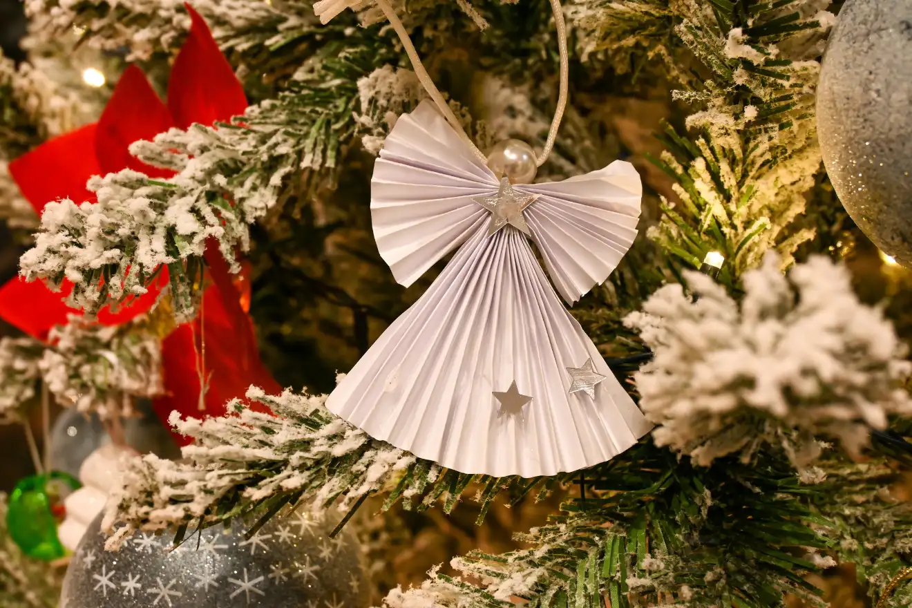 Paper angel ornament with star cutouts hanging on a snow-dusted Christmas tree branch, surrounded by red and silver decorations.