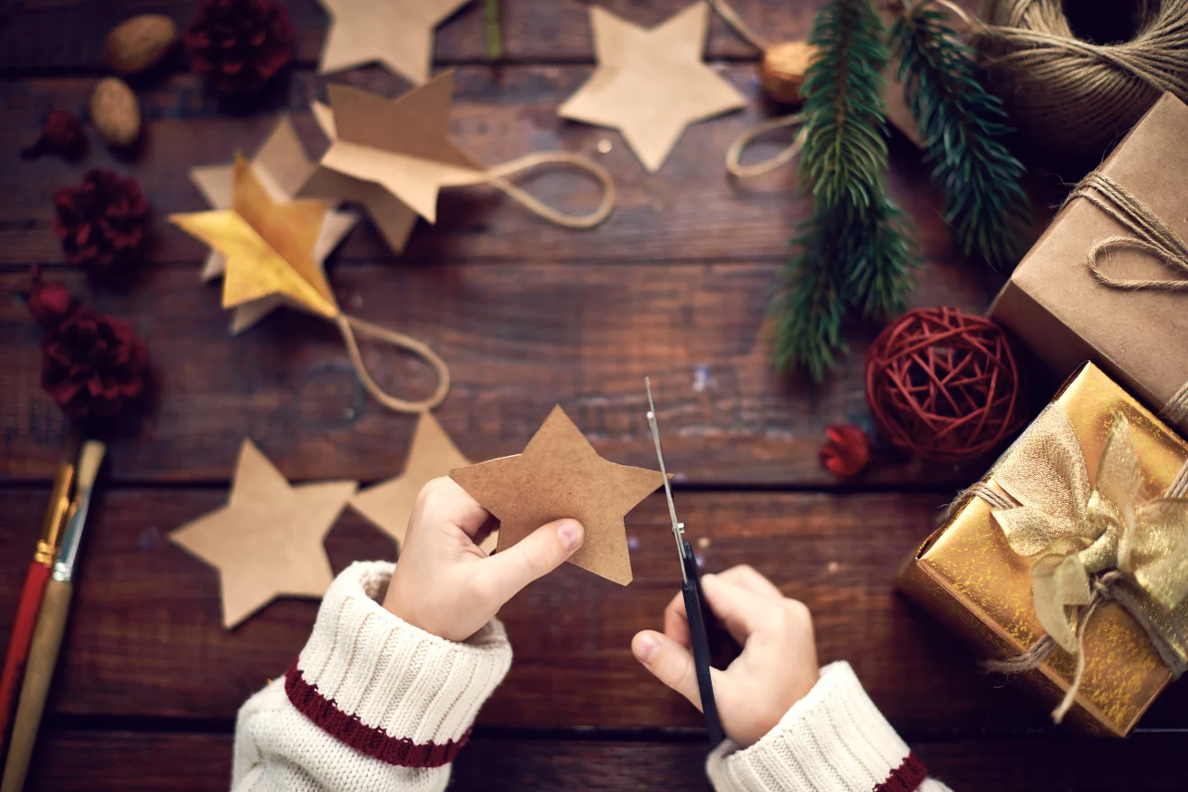 Hands cutting a star shape from brown paper, surrounded by star cutouts, gift boxes, and pine branches on a wooden surface.