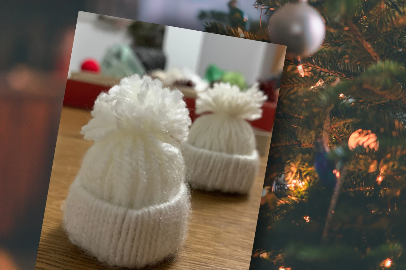 Two white knitted mini hats with pom-poms are placed on a wooden surface, with a blurred Christmas tree to the right.
