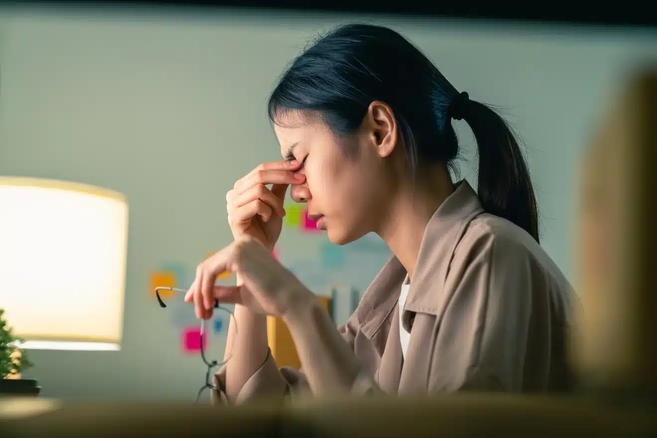 A person sits at a desk, holding their glasses and pinching the bridge of their nose, appearing stressed or tired. A lamp and colorful sticky notes are visible in the background.