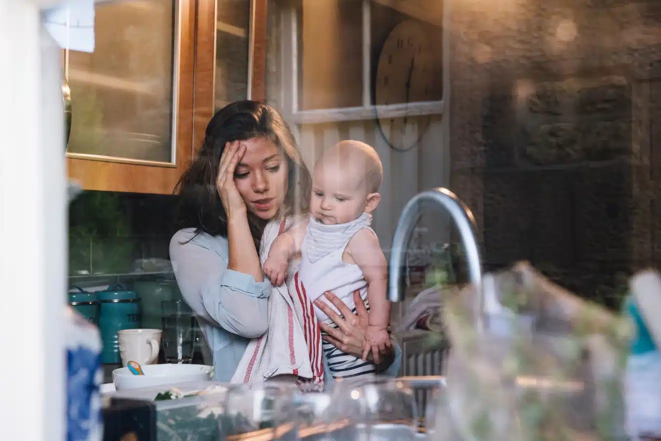 A tired woman holds a baby while standing in a cluttered kitchen, viewed through a window.