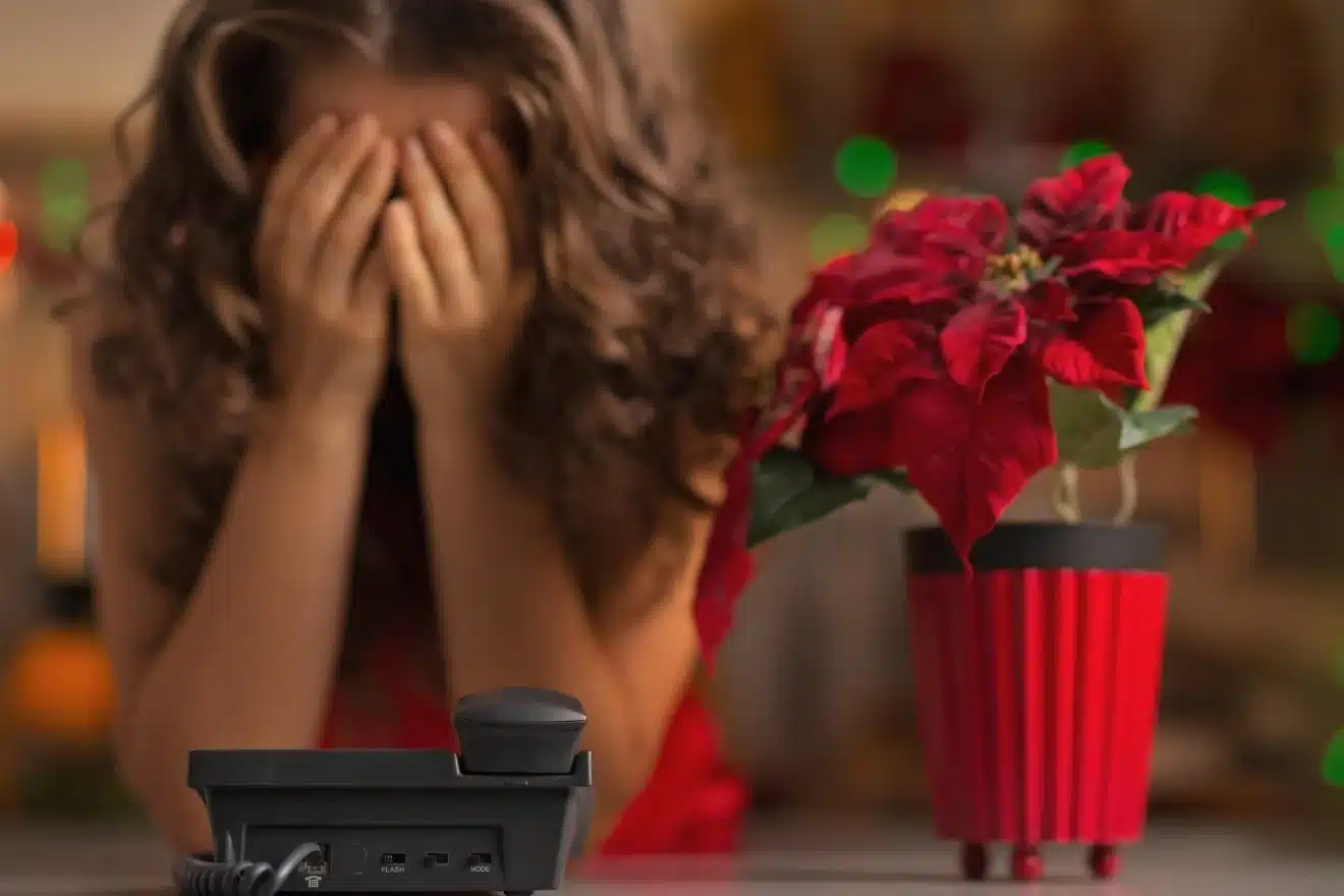 A person, in the midst of managing holiday stress, sits at a table with hands covering their face, accompanied by a phone and a poinsettia plant.
