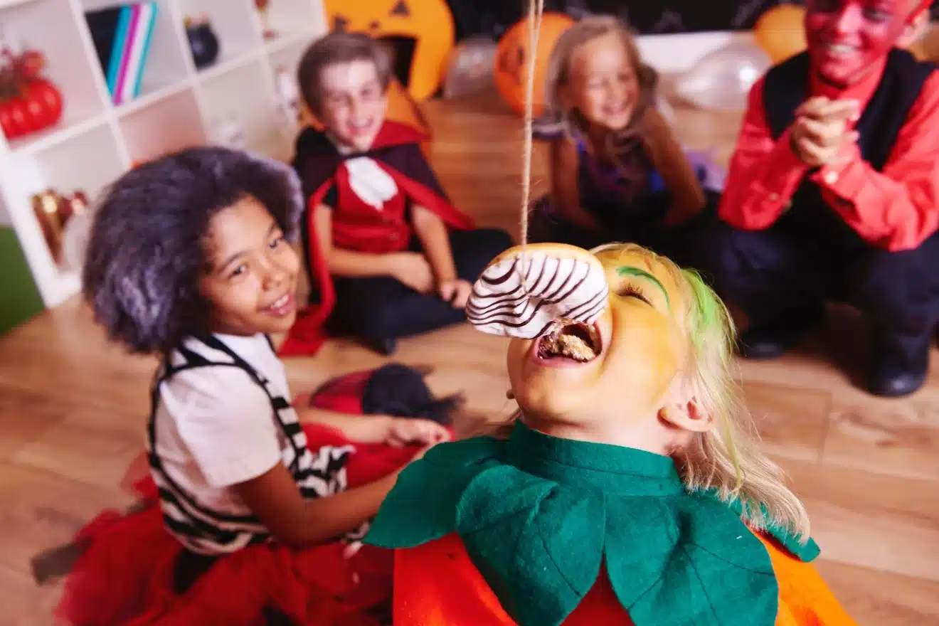 Children in costumes playing a game where one child tries to bite a dangling donut.
