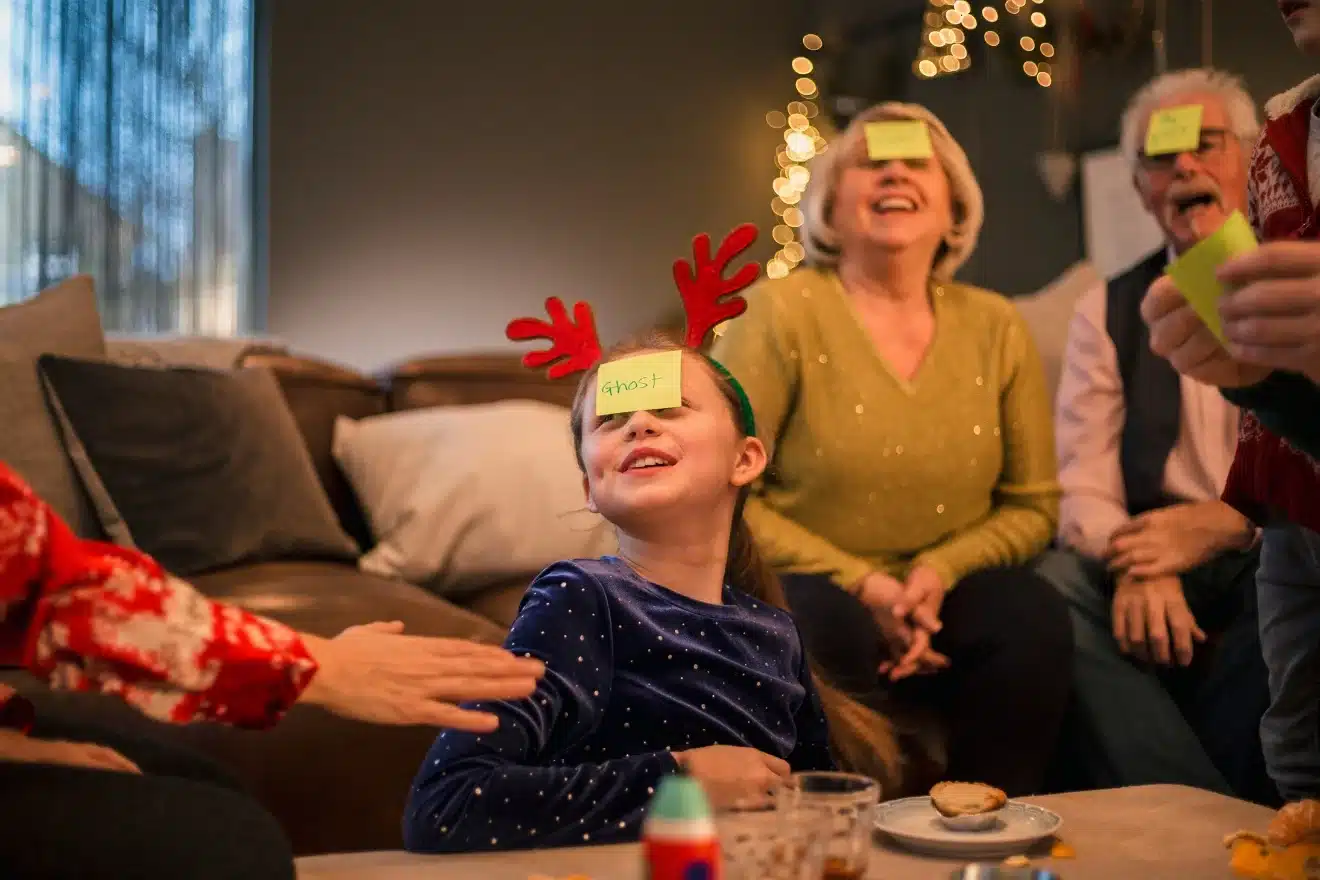 A girl with reindeer antlers and a sticky note on her forehead sits on a couch with others. They are playing a game, with more sticky notes on the foreheads of two other people.