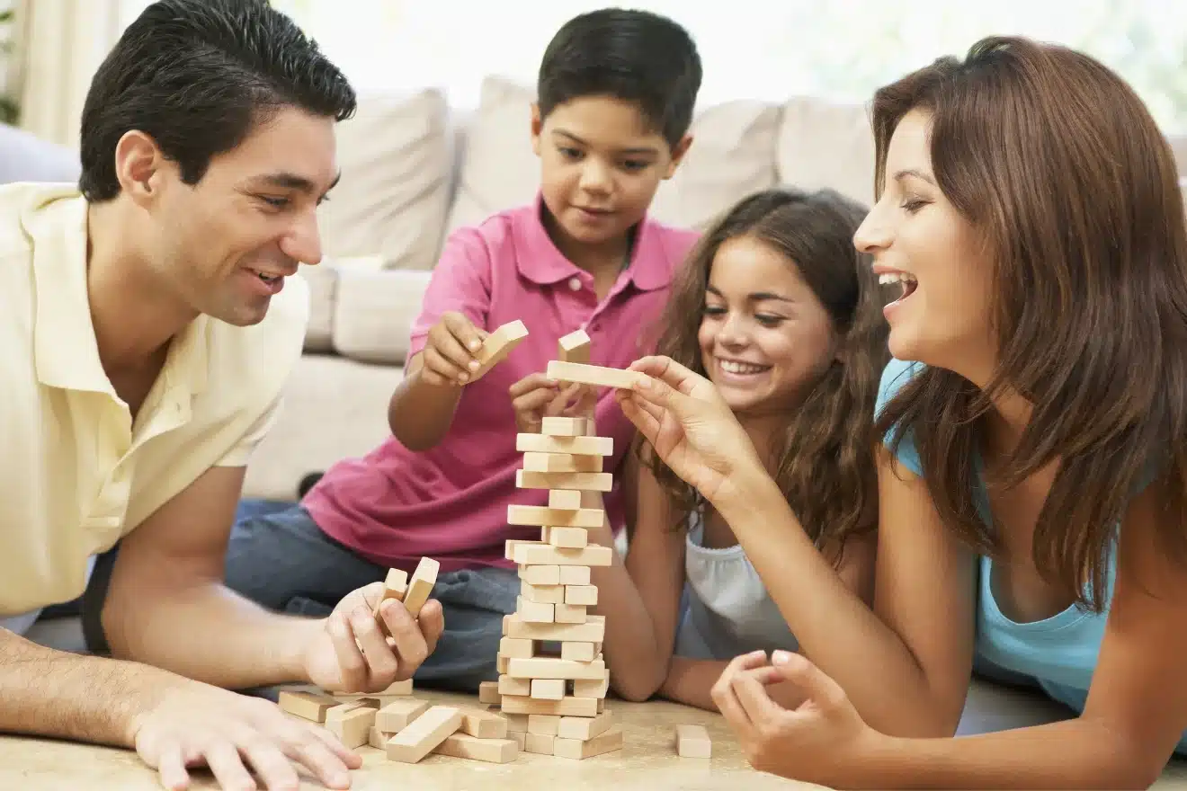 A family of four plays a game of Jenga on the floor, focusing on carefully placing wooden blocks to build a tower.