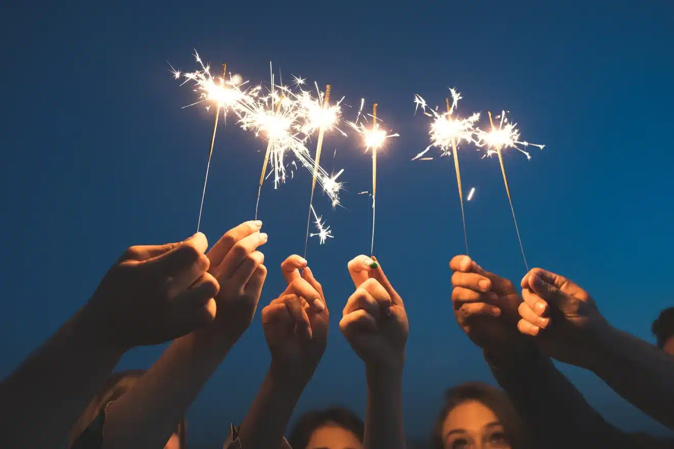 Hands holding lit sparklers against a dark blue sky background.