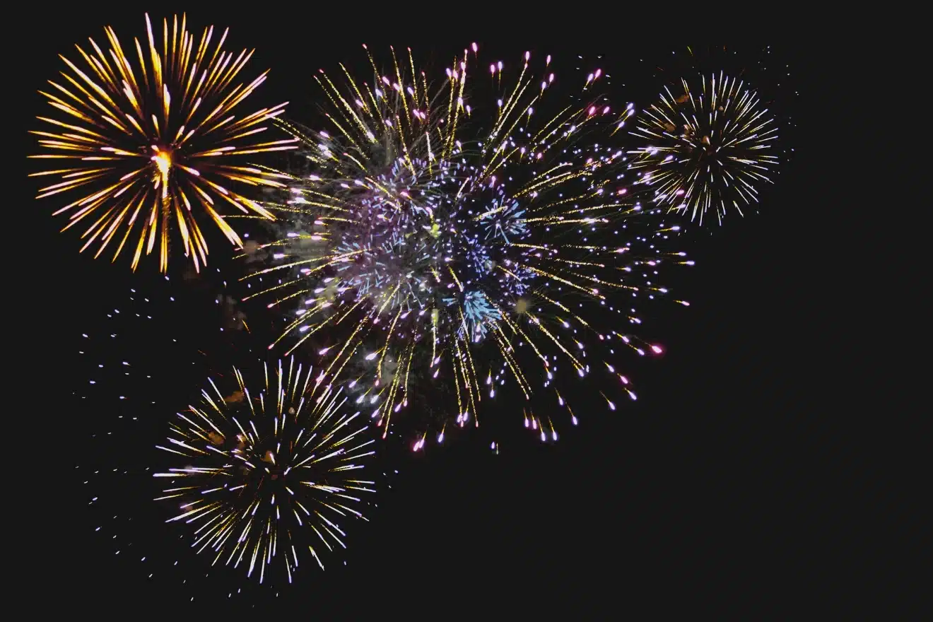 Colorful fireworks burst against a dark sky, displaying various shapes and sizes with bright yellow, purple, and white sparks.