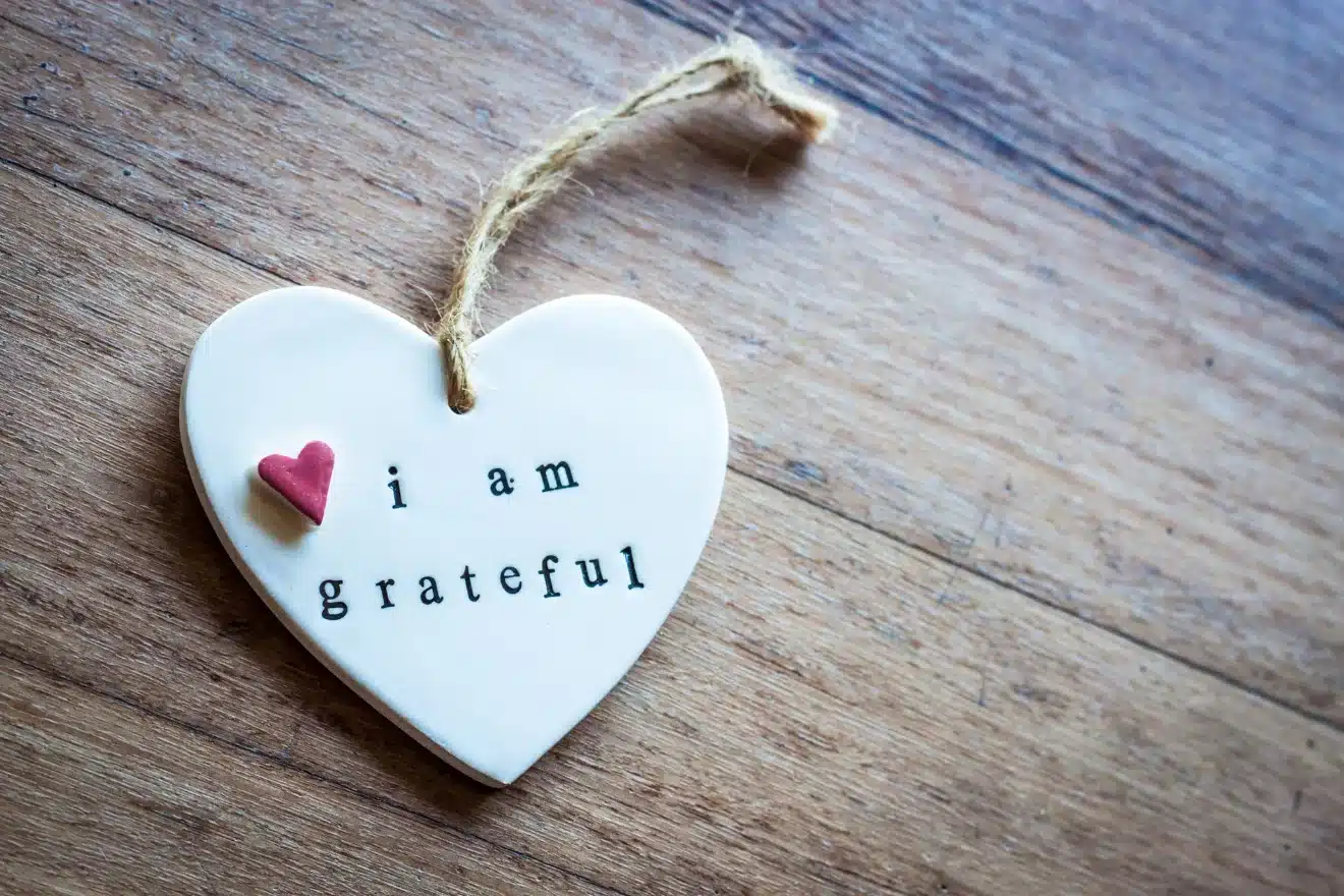 A heart-shaped ornament with the text "i am grateful" and a small red heart is hanging on a wooden surface.