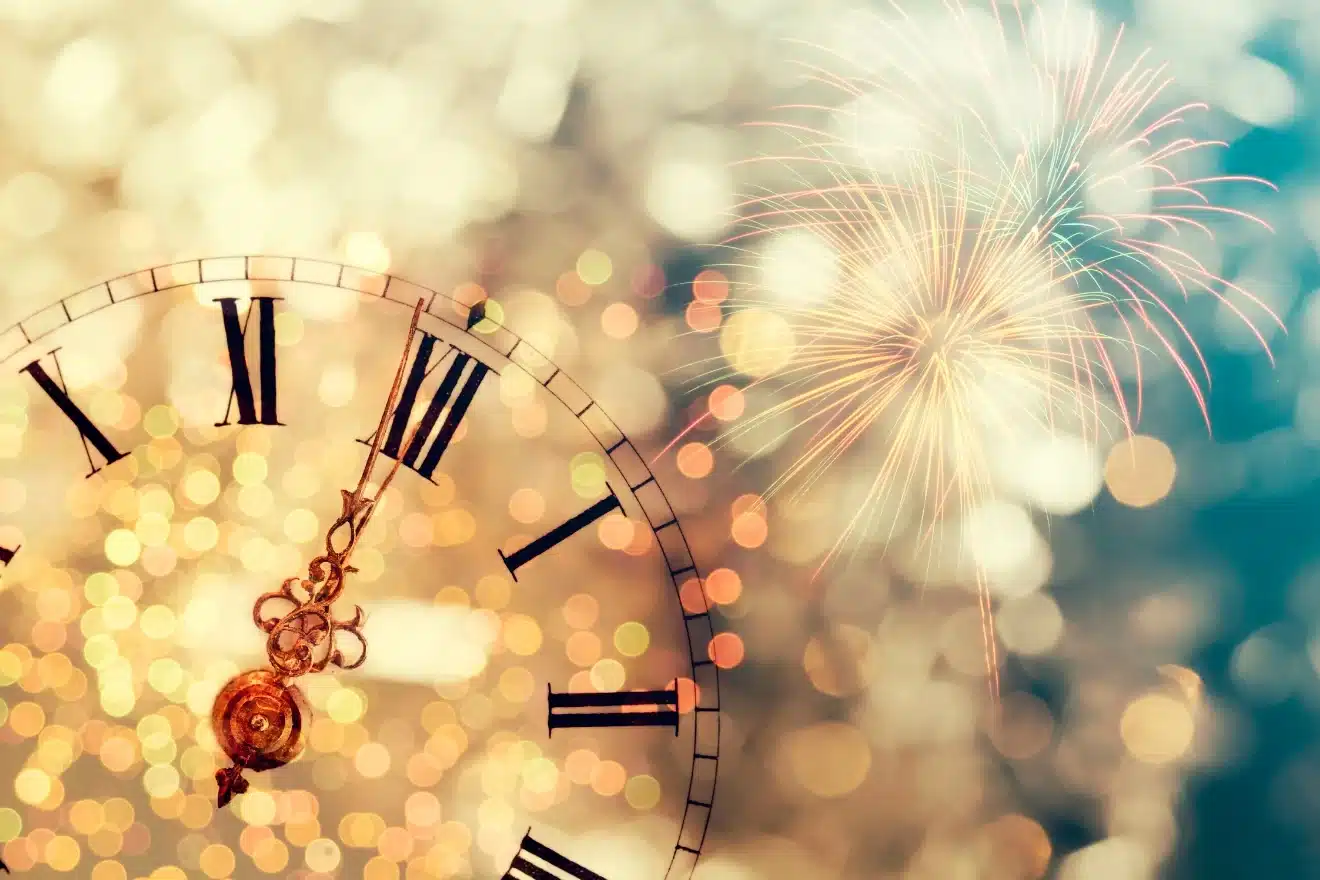 Close-up of a clock showing midnight with Roman numerals, accompanied by blurred lights and fireworks in the background.
