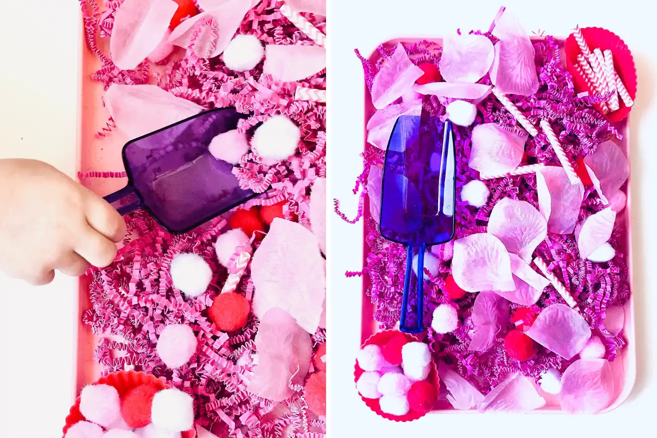 A child's hand uses a purple scoop to move red and white pom-poms and pink shredded paper on a tray filled with similar materials and tissue paper petals.