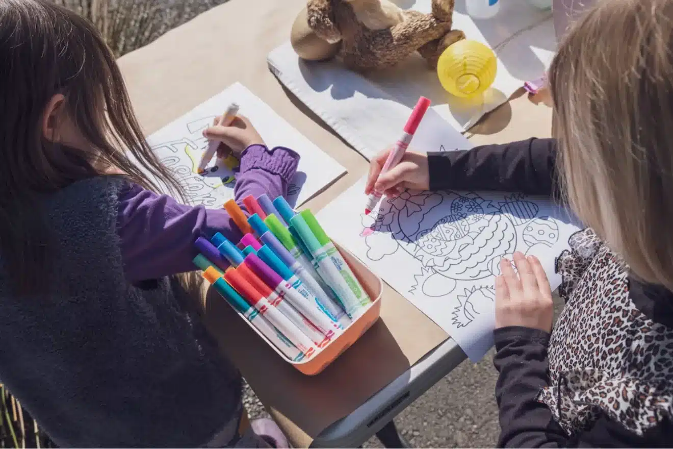 Two children sitting at a table, coloring with markers. A basket of markers is in the center. Coloring pages feature intricate designs. Sunlight casts shadows on the table.